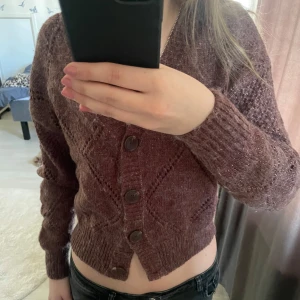 Vinröd/Mörkröd stickad kofta med knappar - Mysig brun stickad kofta med v-ringning och dekorativa knappar framtill. Tröjan har ett diskret rutmönster. Perfekt för lager-på-lager och passar till många olika outfits. 
