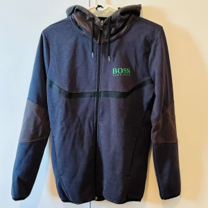 Mörkblå hoodiejacka från Hugo Boss - Säljer en mörkblå hoodiejacka från Hugo Boss med dragkedja och huva. Jackan har svarta detaljer och grönt BOSS-tryck på bröstet. Perfekt för en sportig och avslappnad stil.