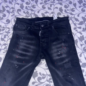 Svarta jeansbyxor från Dsquared2 med färgstänk - Säljer ett par svarta jeansbyxor från Dsquared2 med smal passform. Byxorna har vita och röda färgstänk som ger en cool och unik look. Klassisk femficksmodell med knappgylf och Dsquared2-detalj vid linningen. Endast testade, storlek 170. 