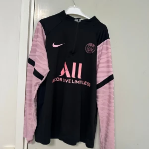 PSG långärmad fotbollströja med rosa detaljer - Säljer en svart långärmad PSG fotbollströja från Nike med rosa detaljer och logga. Tröjan har halv dragkedja framtill, rosa mönstrade ärmar och tryck på bröstet samt ryggen. Perfekt för dig som gillar Paris Saint-Germain. Passar både S och m