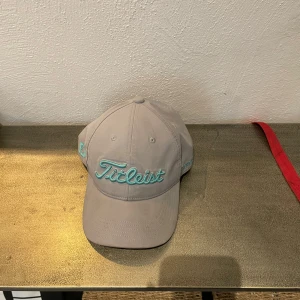 Ljusgrå keps från Titleist - Snygg ljusgrå keps från Titleist med turkos broderad logga framtill och justerbart spänne bak. Kepsen har klassisk form och är perfekt för golf eller vardag. Märke även på sidan och bak. Väldigt fint skick och kommer ny tvättad och fräsch. 