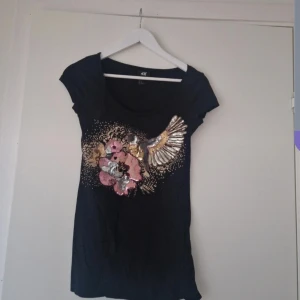 Svart t-shirt med paljettdekor från H&M - Svart t-shirt från H&M med korta ärmar och rund halsringning. Framsidan är dekorerad med stora paljetter i guld, silver och rosa som bildar ett blommotiv och en vinge. Perfekt för dig som gillar att sticka ut!