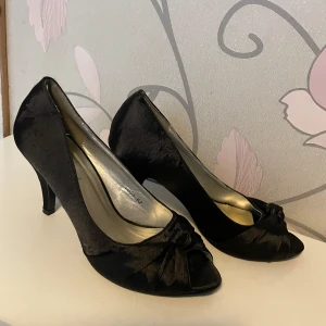 Svarta pumps med rosett och öppen tå - Säljer ett par eleganta svarta pumps från Caprice med öppen tå och fin rosettdetalj framtill. Skorna har en klassisk klack och är tillverkade i ett glansigt syntetmaterial som ger en lyxig känsla. Perfekta för dig som vill ha en stilren och tidlös look.