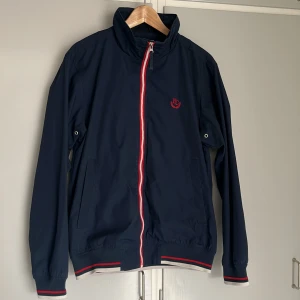 Mörkblå vindjacka från Etirel Small - Säljer en mörkblå vindjacka från Etirel med rödvit dragkedja och randiga detaljer vid ärmslut och midja. Jackan har ståkrage, två sidofickor och rutig insida. Storlek Small.