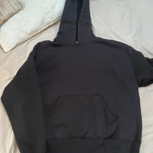 Svart hoodie med ficka framtill - En klassisk svart hoodie med huva och stor magficka. Tröjan har långa ärmar och en avslappnad passform, perfekt för en chill vardagslook. Passar både tjejer och killar som gillar enkel och stilren streetwear.