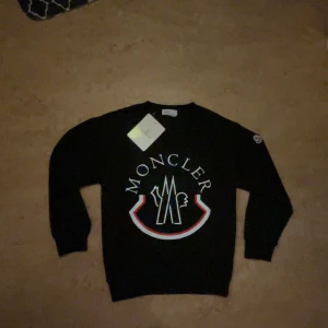 Svart Moncler hoodie med stor logga - Svart sweatshirt från Moncler med stor vit och röd logga framtill. Rund halsringning och långa ärmar. Klassisk och stilren design med Moncler-märke på ärmen. Skriv för mer frågor 