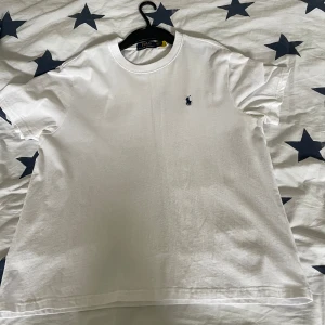 Vit t-shirt från Ralph Lauren - Klassisk vit t-shirt från Ralph Lauren med liten blå broderad logga på bröstet. T-shirten har rund halsringning och korta ärmar. Perfekt basic-plagg att ha i garderoben. Sitter som en M-L