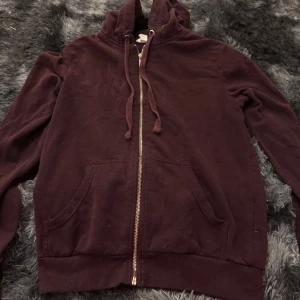 Vinröd hoodie med dragkedja - Mysig vinröd hoodie med dragkedja framtill och två fickor. Tröjan har huva med dragsko och ribbade muddar vid ärmslut och nederkant. Perfekt för kyliga dagar.