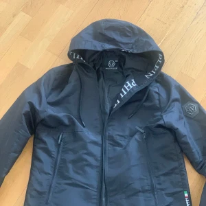Svart vindjacka från Philipp Plein - Snygg svart vindjacka från Philipp Plein med huva och dragkedja. Jackan har två fickor med dragkedja och tydliga logodetaljer på ärmen och insidan. Perfekt för dig som vill ha en stilren och exklusiv look, priset är diskuterbart☺️