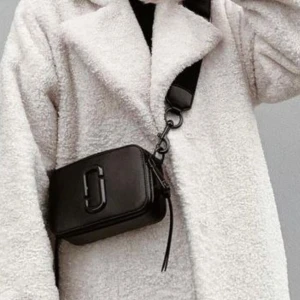 Svart axelväska från Marc Jacobs - Säljer en svart axelväska från Marc Jacobs med bred justerbar axelrem och dekorativ metallogo på framsidan. Väskan har dragkedja upptill och är tillverkad i slätt skinn. Perfekt för att bära det viktigaste med stil. Nypris ca 4100kr dustbag medföljer 