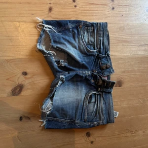 Mörkblå jeansshorts med slitningar - Säljer ett par mörkblå jeansshorts med kraftiga slitningar och fransiga kanter. Shortsen har klassiska fem fickor, bälteshällor och knäppning fram. Perfekta för en avslappnad och trendig look.