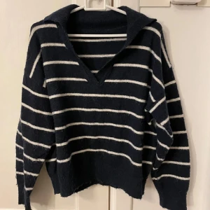 Marinblå stickad tröja med vita ränder - Mysig marinblå stickad tröja med vita horisontella ränder och v-ringad krage. Tröjan har långa ärmar och en avslappnad passform, perfekt för kyliga dagar - kanske en sommarkväll?