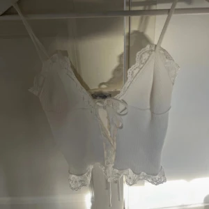 Vit ribbad linnetopp med spetsdetaljer - Säljer en söt vit ribbad linnetopp med spetskant och rosett framtill. Toppen har smala axelband och v-ringning, samt fina spetsdetaljer längs kanterna. Perfekt för sommardagar eller som layering-plagg.