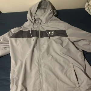 Ljusgrå vindjacka från Under Armour - Snygg ljusgrå vindjacka med huva och dragkedja från Under Armour. Jackan har en mörkare grå panel över bröstet med logga och två sidofickor. Perfekt för blåsiga dagar.
