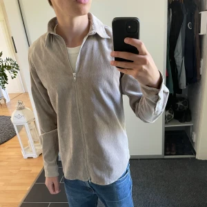 Beige overshirt med dragkedja från Ciszere - Säljer en stilren beige overshirt från Ciszere med dragkedja framtill. Skjortan har klassisk krage och långa ärmar, tillverkad i mjuk manchester. Perfekt att bära öppen eller stängd över en t-shirt. Nyskick då den är oanvänd.