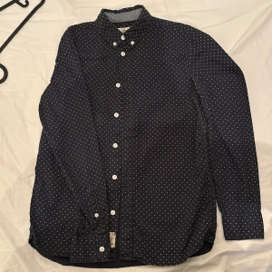 Mörkblå prickig skjorta från L.O.G.G. - Snygg mörkblå skjorta från L.O.G.G. med vita prickar och broderad detalj på bröstet. Skjortan har långa ärmar och klassisk krage, samt knäppning framtill. Perfekt för dig som gillar stilrena mönster!