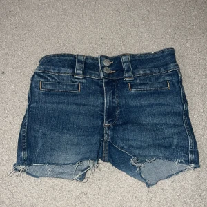 Blå jeansshorts från H&M - Snygga blå jeansshorts från H&M med låg midja och råa, fransiga benslut. Shortsen har två knappar framtill, klassiska fickor både fram och bak samt slitna detaljer för en cool look.