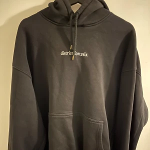 Svart hoodie från District Fortysix - Säljer en svart hoodie från District Fortysix med broderad logga framtill och stor grafisk design på ryggen. Tröjan har huva med dragsko och en stor magficka. Perfekt för dig som gillar streetwear.
