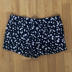 Svarta blommiga shorts från Cotton On - Säljer ett par svarta shorts från Cotton On med vitt och rosa blommönster. De har normal passform och är perfekta för varma dagar. Stängs med dragkedja och knapp framtill. Superfina till sommaren!