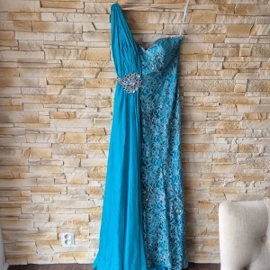 Turkos långklänning från Terani Couture - Säljer en turkos långklänning från Terani Couture med en axel och vacker spetsdetalj. Klänningen har en glittrig broderad dekoration i midjan och är tillverkad i polyester. Perfekt för bal eller festliga tillfällen.