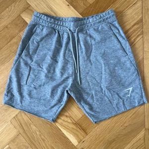 Grå shorts från Gymshark - Lätta grå shorts från Gymshark med snörning i midjan och diskret logga på vänster ben. Perfekta för träning eller chill, med två sidofickor och mjukt material som känns skönt mot huden.