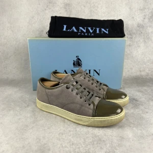 Grå sneakers från Lanvin med lackad tå - Säljer ett par grå sneakers från Lanvin med snygg kontrast i form av mörkgrå lackad tå och matchande snörning. Skorna har en beige sula och är tillverkade i mocka och skinn. Perfekta för dig som gillar stilrena detaljer.