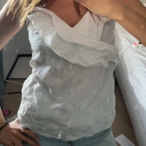 Vit ärmlös blus med volang - Säljer en söt vit ärmlös blus med broderade blommor och volangdetalj framtill. Perfekt till sommaren och passar till både jeans och kjol. Blusen har en luftig och feminin design.
