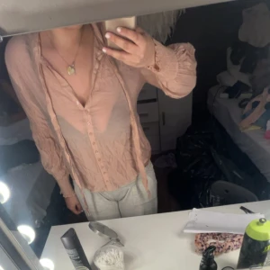 Ljusrosa blus med knytband - Säljer en ljusrosa blus med knappar framtill och långa ärmar. Blusen har knytband vid halsen och är tillverkad i ett tunt, lätt material som ger en luftig känsla. Perfekt för dig som gillar romantiska detaljer.