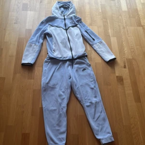 Nike tech fleece - Säljer en snygg Nike tech fleece. Hoodien i ljusblått och vitt med svarta detaljer och dragkedja. Hoodien har huva, lång ärm och en modern design med Nike-logga på bröstet. Byxorna är ljusblå och de är i storlek 158-170cm men passar xs. Perfekt för dig som gillar sportig stil.
