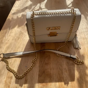 Vit axelväska från Guess med guldkedja - Säljer en stilren vit axelväska från Guess med guldfärgad kedja och logga framtill. Väskan har en strukturerad yta, dekorativ tofs och ett elegant spänne. Perfekt för dig som vill ha en snygg accessoar med lyxig känsla.