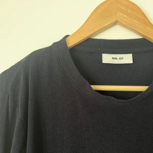 Mörk marinblå långärmad tröja från NN.07 - En stilren svart långärmad tröja från NN.07 med rund halsringning. Tröjan är tillverkad i ett mjukt, strukturerat material som ger en avslappnad men ändå snygg look. Perfekt för lager-på-lager eller som den är.
