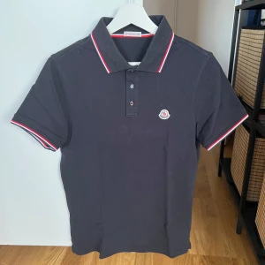 Mörkblå pikétröja från Moncler - Säljer en mörkblå pikétröja från Moncler med klassisk krage och knappar framtill. Tröjan har röda och vita detaljer på krage och ärmslut samt Moncler-logga på bröstet. Perfekt för en stilren och sportig look.