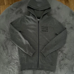 Grå zip hoodie från Peak Performance - Säljer en grå zip hoodie från Peak Performance med dragkedja framtill och svart logga på bröstet. Tröjan har huva och långa ärmar, perfekt för en avslappnad stil. Mjuk och bekväm med klassisk passform.
