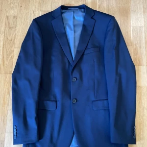 Mörkblå kavaj från Dressmann - Säljer en stilren mörkblå kavaj från Dressmann i storlek 48L (slim fit). Kavajen har två knappar framtill, fickor med lock och en bröstficka. Fodret är ljusblått med röda detaljer. Perfekt för dig som vill ha en klassisk look. Kavajen är endast använd 1 gång. Nytt skick inga konstigheter.