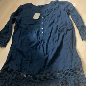 Svart klänning med spetsdetaljer från Vero Moda - Svart klänning från Vero Moda med långa ärmar och fina spetsdetaljer vid ärmslut och nederkant. Knappstängning framtill och rund halsringning. Perfekt för dig som gillar stilrena plagg med extra detaljer.