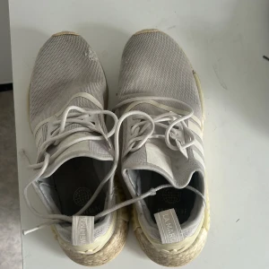 Adidas NMD R1 sneakers i ljusgrått - Säljer ett par Adidas NMD R1 sneakers i ljusgrått med vita detaljer och klassiska tre ränder på sidan. Skorna har snörning, platt sula och är tillverkade i syntetmaterial och textil. Perfekta för en sportig och stilren look.