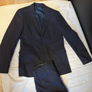 Mörkblå kostym  - Klassisk mörkblå kostym från Riley med kavaj och matchande byxor. Kavajen har två knappar, smala slag och fickor med lock. Byxorna har enkel design med knapp och dragkedja. Perfekt för formella tillfällen.