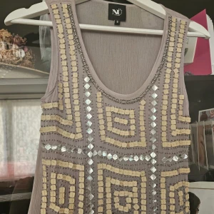 Beige och silvermönstrad klänning från NÜ - Säljer en ärmlös klänning från NÜ i beige med geometriska mönster av silverfärgade paljetter och pärlor. Klänningen har rund halsringning och är i ett lätt, transparent material. Perfekt för dig som vill sticka ut med snygga detaljer.
