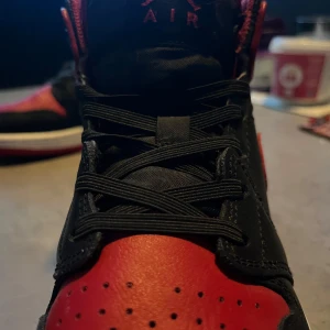 Nike Air Jordan 1 Retro High OG Bred - Säljer ett par klassiska Nike Air Jordan 1 Retro High OG i svart och rött med vita sulor. Skorna har snörning, rund tå och är tillverkade i skinn. Perfekta för dig som vill sticka ut med en ikonisk sneaker.