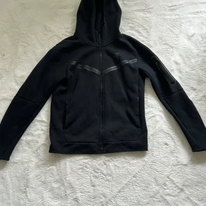 Svart hoodiejacka med dragkedja - Svart hoodiejacka med hel dragkedja framtill och huva. Jackan har diskreta detaljer på bröstet och en ficka med dragkedja på ena ärmen. Perfekt för en sportig och avslappnad stil.