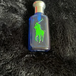 Blå parfymflaska från Ralph Lauren Polo - Säljer en snygg 75% full parfymflaska från Ralph Lauren Polo. Flaskan är blå med ett stort grönt polospelar-tryck på framsidan och har ett silvrigt lock. Perfekt för dig som gillar klassiska och sportiga dofter.