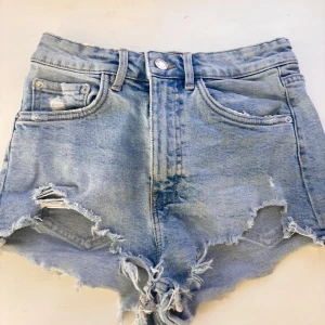 Ljusblå jeansshorts med fransar  - Säljer ett par ljusblå jeansshorts med hög midja och råa, fransiga kanter. Shortsen har slitningar framtill och klassiska fem fickor. Perfekta för varma dagar och en avslappnad stil.