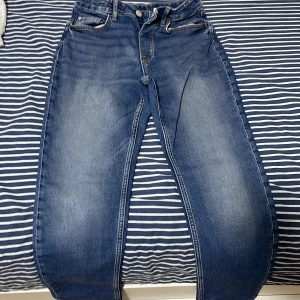 Blå jeans från H&M med normal passform - Säljer ett par klassiska blå jeans från H&M med normal passform och raka ben. Jeansen har fem fickor, dragkedja och knapp framtill samt bälteshällor. Perfekta till vardags och enkla att matcha med olika stilar.