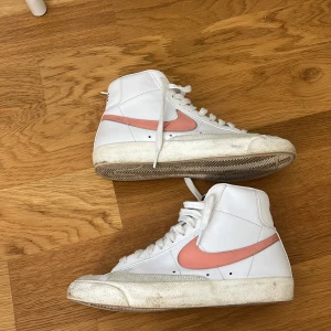 Nike Blazer Mid sneakers med rosa swoosh - Säljer ett par vita Nike Blazer Mid sneakers med hög modell och rosa swoosh på sidorna. Skorna har snörning framtill, rund tå och platt sula. Klassisk Nike-logga på plös och häl. Perfekta för dig som gillar retrostil och streetwear. Pris går att diskutera. Smuts på sulan går att tvätta bort.