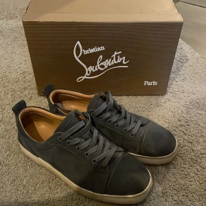 Mörkblåa sneakers från Christian Louboutin - Säljer ett par grå sneakers från Christian Louboutin med klassisk röd sula. Skorna har snörning, rund tå och vit platt sula. Insidan är beige och materialet känns lyxigt och robust. Perfekta för dig som vill ha något stilrent men ändå exklusivt.