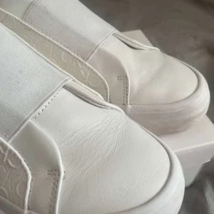 Vita sneakers från Calvin Klein - Säljer ett par stilrena vita sneakers från Calvin Klein med präglad CK-logga runtom och bred resår över vristen. Skorna har en chunky sula och är helt vita, vilket ger en clean och modern look. Perfekta för dig som gillar minimalistisk stil.