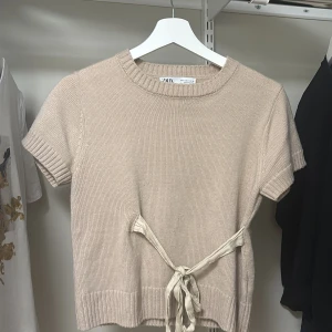 Beige stickad topp med knyt från Zara - Säljer en beige stickad topp från Zara med korta ärmar och knytband i midjan. Tröjan har rund halsringning och ribbade kanter. Perfekt för en stilren och enkel look🤗🤗