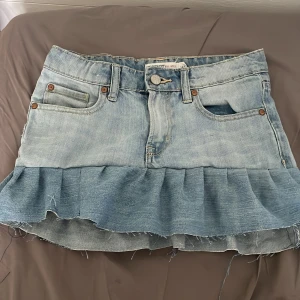 Ljusblå jeanskjol med volang - Säljer en ljusblå jeanskjol med volangkant nedtill. Kjolen har klassiska jeansfickor fram och bak, bälteshällor och knäppning framtill. 