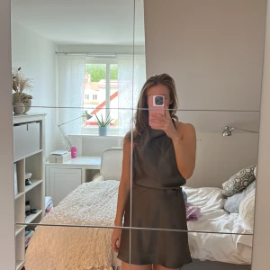 Mörkgrå satin klänning med öppen rygg från zara  - Säljer en mörkgrå kort klänning i satin med öppen rygg och knytband vid axlarna. Klänningen har en slät, glansig yta och en enkel, elegant design som ger en stilren look.