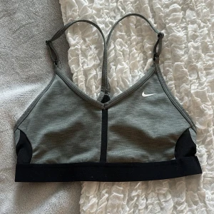 Grå sport-bh från Nike - Sport-bh från Nike i grått med svarta detaljer och justerbara axelband. Liten vit Nike-logga framtill. Perfekt för träning och har en tight passform med bred resår nedtill.